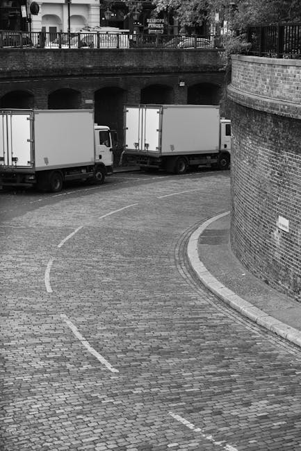 A black-and-white photograph showing three medium-sized box trucks parked in a curved, cobblestone loading area adjacent to a brick wall and an archway under a raised walkway. The trucks are positione