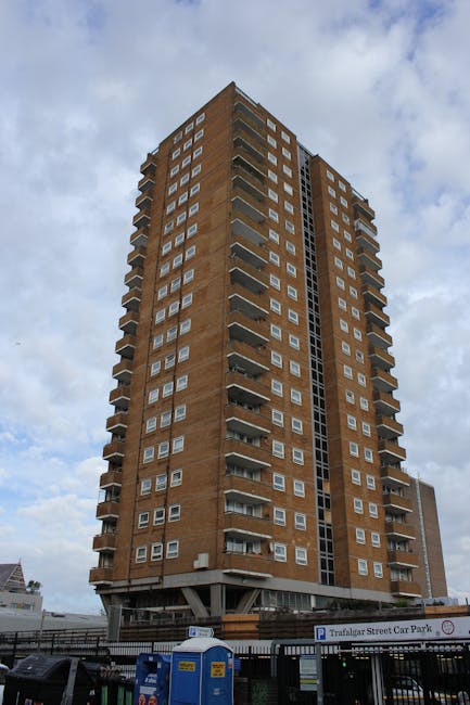 A tall, multi-storey residential building with a brick exterior and numerous balconies, situated in an urban area under a partly cloudy sky. The building features a modern design with symmetrical wind